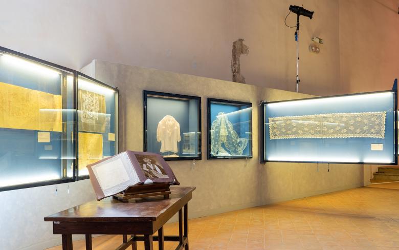 Immagine: Display cases containing tulle artifacts and historical documents. In the foreground, an open book on a table with a photograph. 